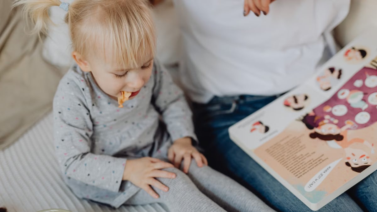 Cute toddler with ponytails enjoying a snack next to a book and parent. Cozy indoor scene.